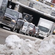 Des voitures et des camions circulent dans une rue enneigée après être passés sous un viaduc, tandis qu'on voit un banc de neige à l'avant-plan.