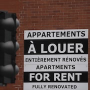 Une pancarte disant «Appartements à louer, entièrement rénovés, Apartments for rent, fully renovated» sur la façade en briques d'un immeuble, tandis qu'on voit un feu de circulation devant.