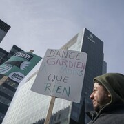 Un homme tient une pancarte sur laquelle on peut lire : « D'ange gardien à moins que rien ».