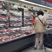 Une femme avec un panier se tient devant le comptoir des viandes dans un supermarché.
