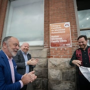 Trois personnes sourient et applaudissent devant une enseigne sur un bâtiment qui porte la mention «Clinique familiale de Montréal Autochtone».