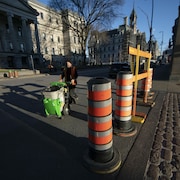Un employé municipal pousse un bac sur roulettes derrière une rangée de cônes orange bloquant le passage dans une rue du Vieux-Montréal.
