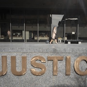 Une femme tirant une petite valise sur roulettes marche devant le palais de justice de Montréal avec à l'avant-plan le logo « Justice ».