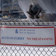 Une pancarte «Port de Montréal - Accès interdit - No trespassing» sur une clôture derrière laquelle on aperçoit l'ancre d'un bateau et, au loin, une remorque transportant des voitures.