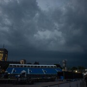 Vue lointaine d'une scène de concert et de gradins bleus sur les plaines d'Abraham à Québec, sous un ciel couvert et sombre. Au premier plan, le terrain est dégagé, avec quelques silhouettes de personnes visibles. Des bâtiments aux lumières allumées sont discernables en arrière-plan, sous un ciel chargé de nuages orageux. L'image capture le moment juste avant une perturbation météorologique majeure.