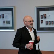 Yves Frenette, l'air sérieux, marche dans un corridor avec un café à la main.