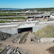 Vue aérienne d'un chantier routier où un pont en béton en construction surplombe une voie ferrée près d'un cours d'eau.