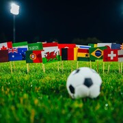 Un ballon de soccer est posé devant une multitude de drapeaux internationaux piqués sur une pelouse.