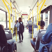 Une adolescente est debout dans l'allée pendant les trajets en autobus. D'autres adultes sont assis de dos, le regard vers l'avant du véhicule.