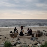 Des gens sont sur une plage, assis sur des bûches de bois en cercle.