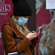 Une femme coiffée d'une tuque en laine et le visage recouvert d'un masque médical est penchée sur son téléphone cellulaire.
