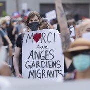 Une femme tenant une pancarte sur laquelle il est inscrit : « Merci aux anges gardiens migrants ».