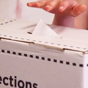 Gros plan d'une boite de scrutin, main mettant bulletin de vote dans la boite.