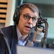 Un homme, casque d'écoute sur la tête, parle dans le micro d'un studio de radio. 