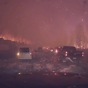 Des voitures qui roulent la nuit sur une route sous une pluie de braises d'un immense incendie tout proche.