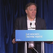Le premier ministre du Québec parle pendant une conférence de presse derrière un lutrin qui porte la mention « Le hockey, notre fierté ».