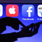 Une personne utilise un téléphone cellulaire devant un écran affichant des logos de Google, Apple, Facebook et Amazon.