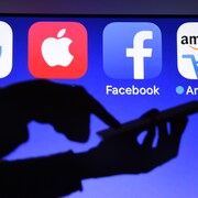 Une personne utilise un téléphone cellulaire devant un écran affichant des logos de Google, Apple, Facebook et Amazon.