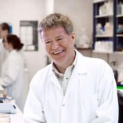 Un homme souriant et portant un sarrau blanc se trouve dans un laboratoire de recherche scientifique.
