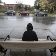 Un jeune homme assis seul de dos sur un banc en bois, dans une rue, sous la pluie.