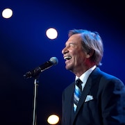 Jean-Pierre Ferland, debout sur scène devant un micro, sourit en chantant.