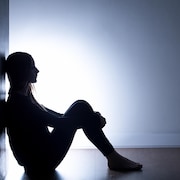 Une jeune femme est assise par terre, adossée contre un mur dans l'obscurité.