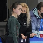 La jeune patineuse regarde la glace d'un air inquiet, pendant que son entraîneuse lui parle, accotée sur la bande de la patinoire. Un homme du Comité olympique russe, non identifié sur la photo, se trouve à leur gauche. 