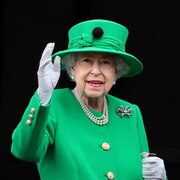 La reine, vêtue de vert, salue les gens avec sa main.