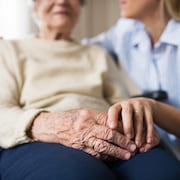 Une personne âgée en fauteuil roulant, à qui une femme plus jeune tient la main.