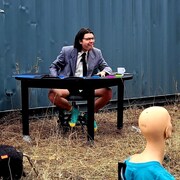 Un comédien est assis derrière une table, devant un conteneur de transport ferroviaire; il porte un complet, mais sans pantalon.