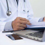 Un médecin avec un stéthoscope autour du cou regarde ses notes dans un cahier en face de son ordinateur portable et de son cellulaire.