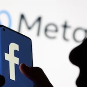La silhouette d'une personne tenant un cellulaire sur lequel apparaît le logo de Facebook, avec le logo de Meta en arrière-plan.