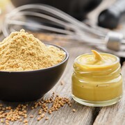 Des graines de moutarde dans une cuillère en bois, de la moutarde en poudre dans un bol et de la moutarde Dijon dans un pot en verre disposés sur une table en bois. 