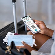 Une femme assise dans un aéroport vérifie son passeport de vaccination de la COVID-19 sur son téléphone, tout en tenant un masque et une carte d'embarquement. 