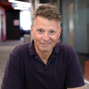 L'homme pose en souriant, assis dans le couloir d'un édifice.