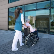 Une jeune femme ramène chez lui un homme âgé en fauteuil roulant.