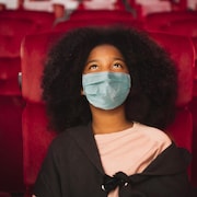 Une jeune fille portant un masque de protection est assise dans une salle de cinéma, avec du maïs soufflé. 