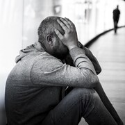 Une homme visiblement triste est assis par terre. Il tient son visage dans ses mains. Il est habillé avec un chandail à capuche et un pantalon en jean.