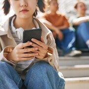Une adolescente regarde son téléphone cellulaire pendant qu'un groupe de jeunes, à l'arrière-plan, semble se moquer d'elle.