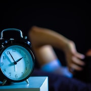 Une personne allongée se tient la tête dans le fond de l'image avec une horloge au premier plan qui indique 1 h 55 du matin. 