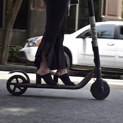 Une femme portant des chaussures à talons conduit une trottinette électrique sur une rue. 