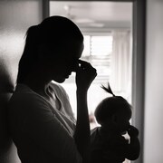 Silhouette d'une femme adossée contre un  dans un couloir et ayant son bébé dans les bras. Elle se touche le front comme si elle avait mal à la tête.