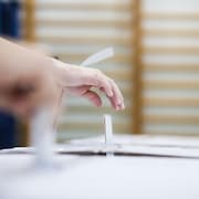 Gros plan sur des mains qui déposent un bulletin de vote dans une urne en carton, dans un gymnase.
