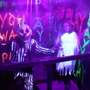 Un clown sinistre et une mariée lugubre fixent la caméra dans une allure effrayante. En arrière-plan, des messages « YOU WANT TO PLAY » ont été peints sur les murs.