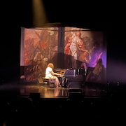 Une femme vêtue tout en blanc joue du piano à queue sur une scène.
