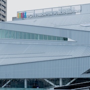 Bâtiment en acier gris très pâle de forme atypique. Plusieurs fenêtres, Lettrage blanc tout en haut disant : Edmonton Public Library. Ciel gris. Juillet 2023.