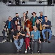 Un groupe de musiciens composé de 10 hommes et une femme sourient en tenant leurs instruments en mains (cuivres, guitare, accordéons, violon, contrebasse).