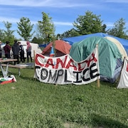 Des tentes sur un terrain. Une banderole, où c'est écrit « Canada complice », est devant.