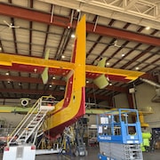 Un avion-citerne est entouré d'échelles dans un hangar. Des travailleurs sont sur le toit de l'avion. 