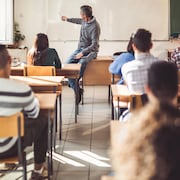 Un enseignant est assis sur son bureau et montre des choses au tableau. Des élèves sont assis en classe et écoutent.